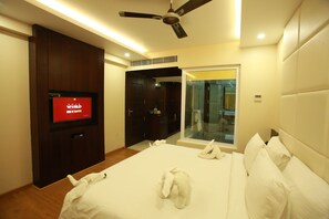 Habitación de hotel con una cama, ventilador de techo, televisor y puerta de vidrio que da a un baño.