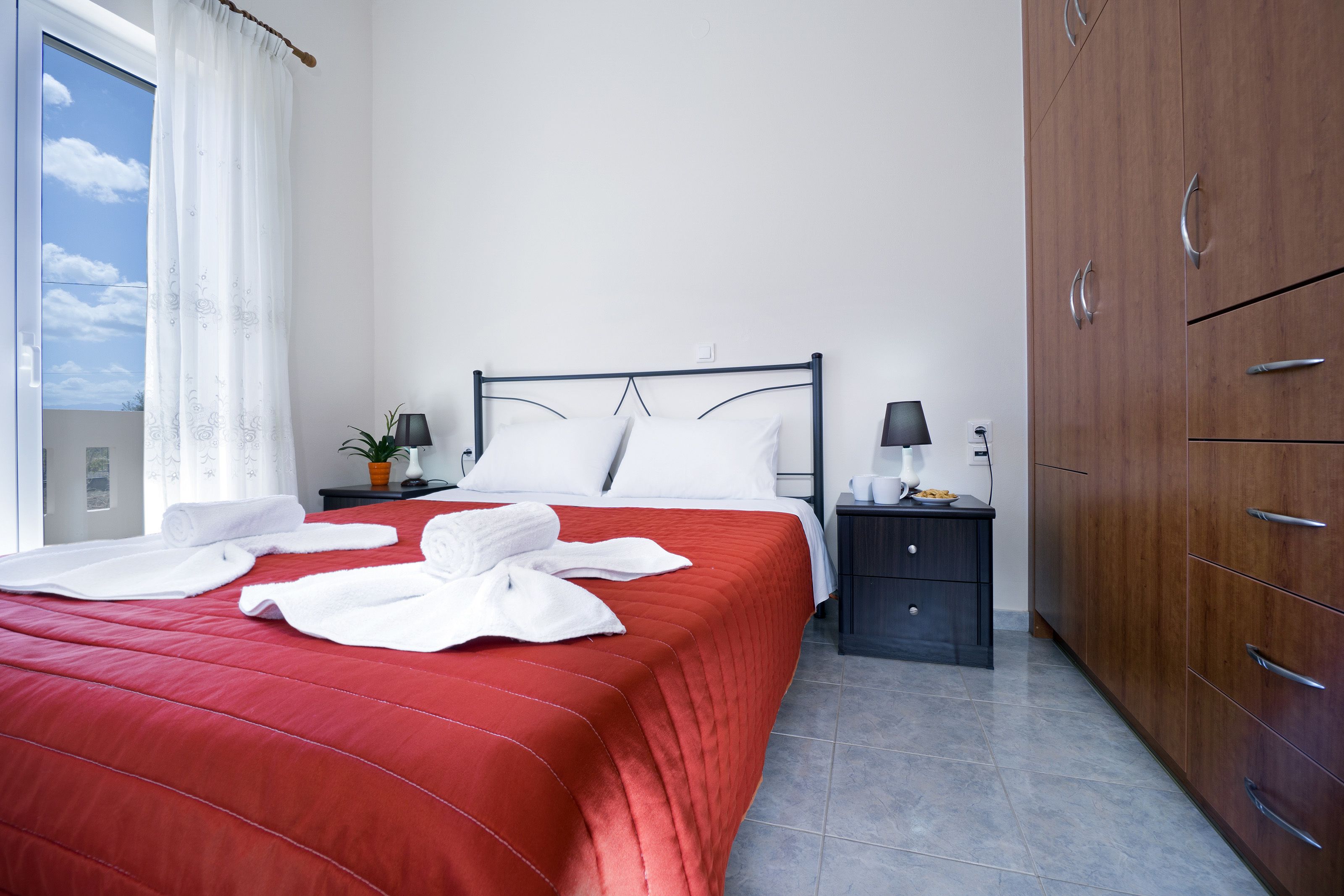 A neatly made bed with white linens and a red bedspread, a bedside table with a lamp, a small potted plant, and a wardrobe with multiple drawers.