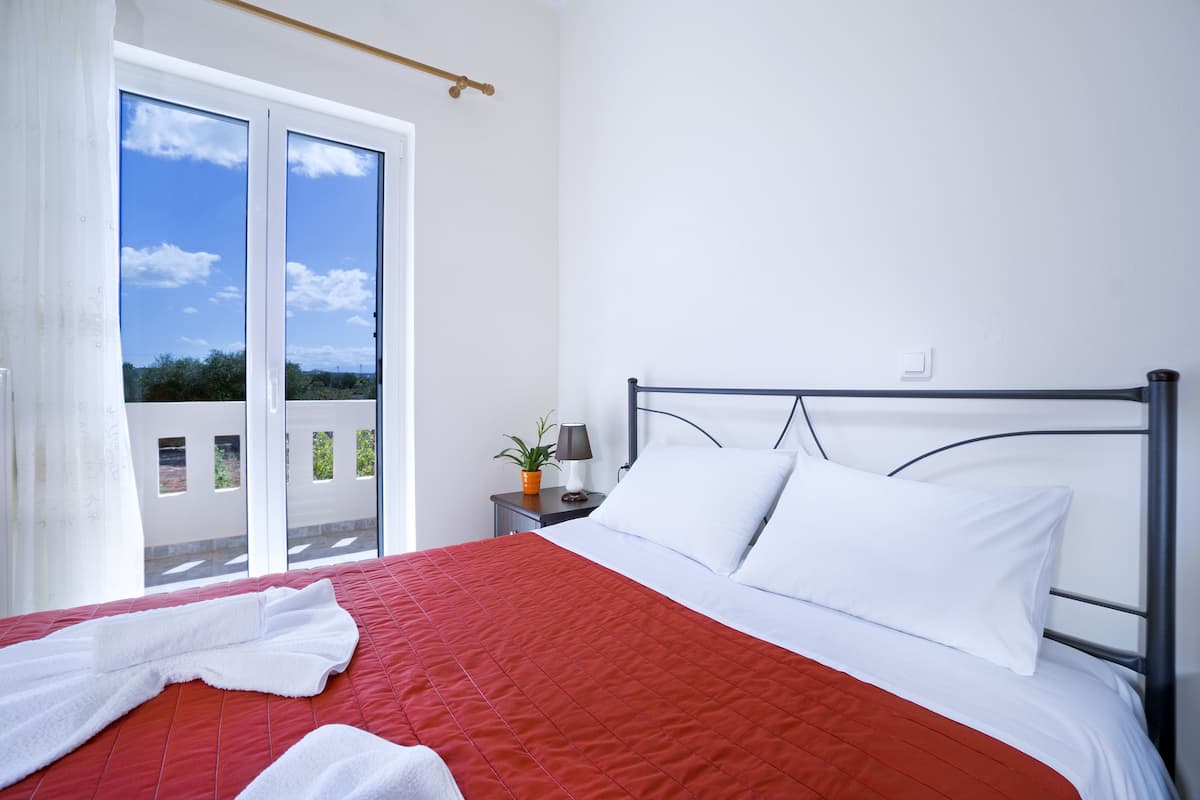 Une chambre d’hôtel avec un lit, des oreillers blancs, une couvre-lit rouge, une table de chevet avec une lampe et une vue sur l’extérieur grâce à une porte coulissante.