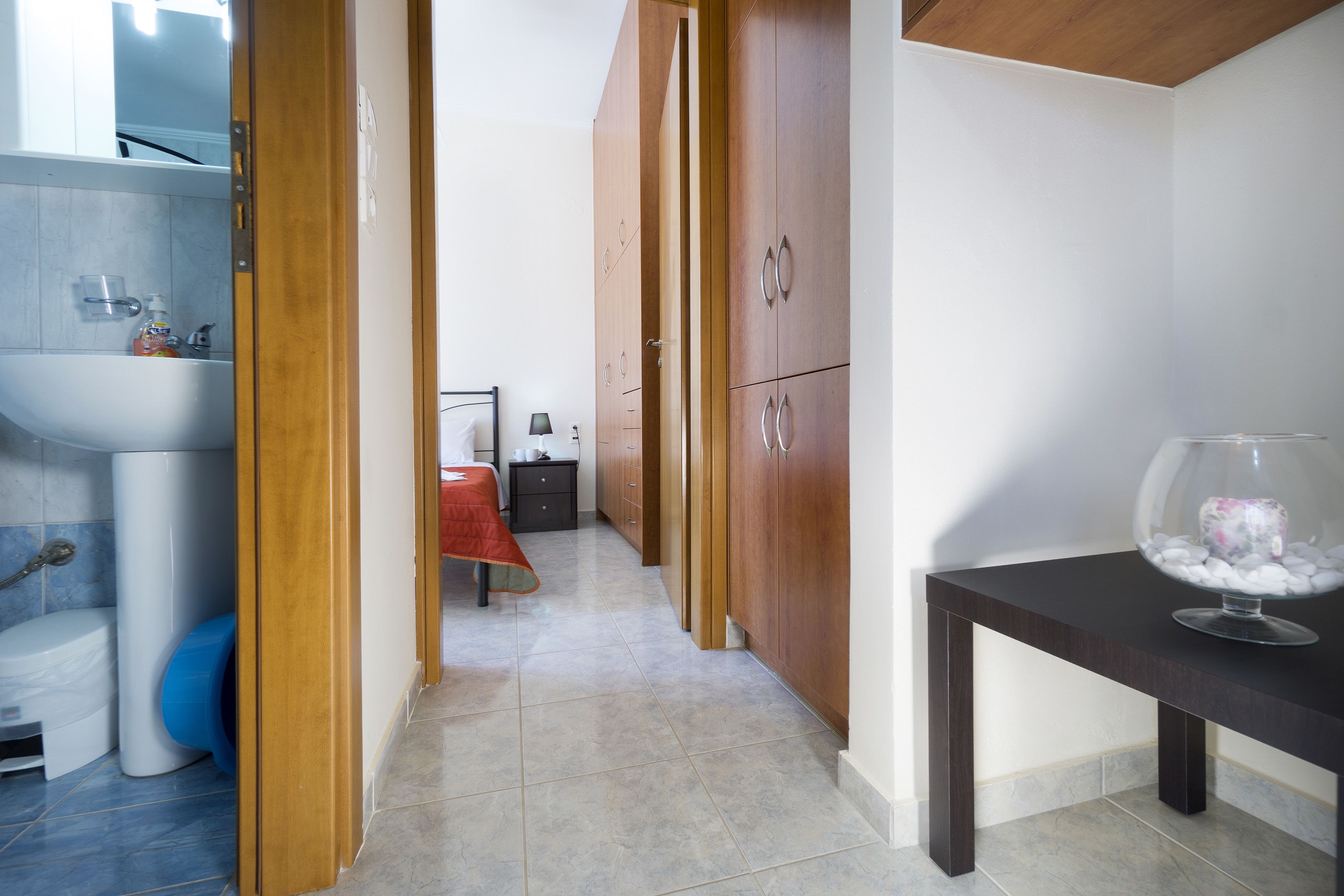 Quarto de hotel com banheiro, uma cama com manta vermelha, um guarda-roupa de madeira e uma pequena mesa com uma tigela de vidro decorativa.