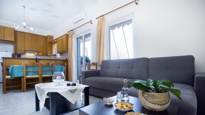 A modern living room with a grey sofa, a dining table with chairs, a kitchen area, and a potted plant. - Skamagkas G. Apartments (Chania)