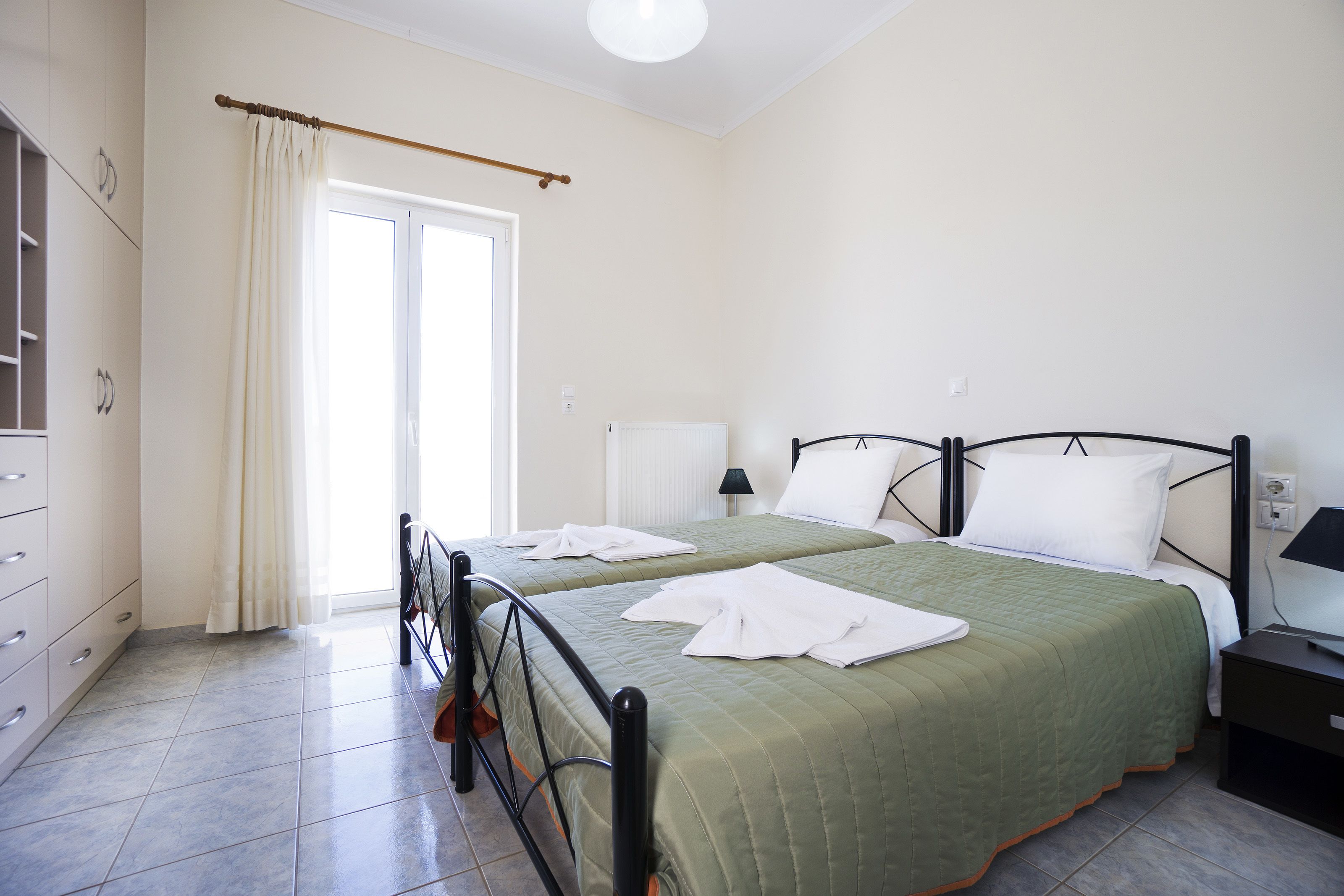 A bedroom with two beds, a wardrobe, a radiator, and a window with curtains.