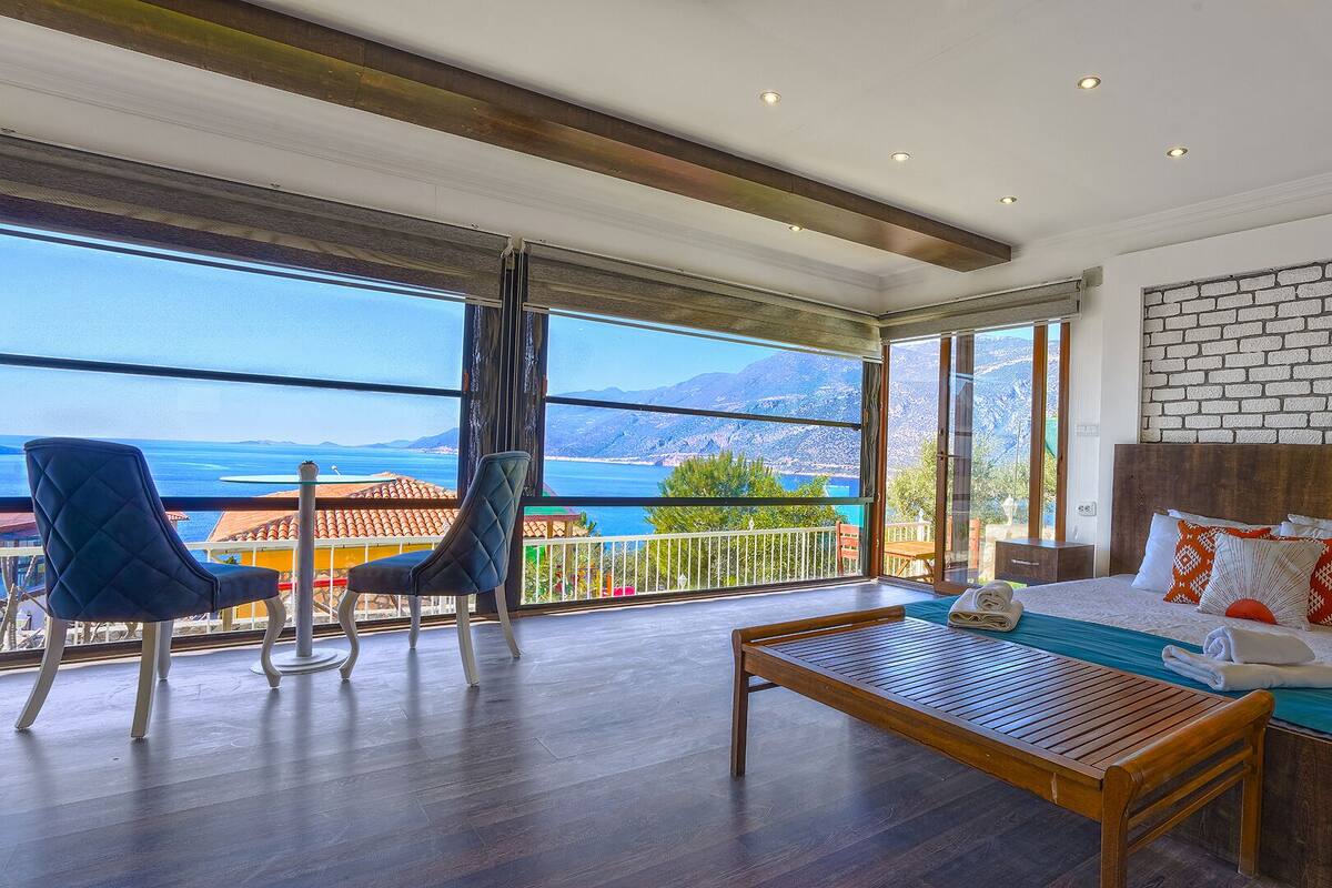 A hotel room with a large balcony, wooden flooring, a bed with pillows, and a view of the sea and mountains.