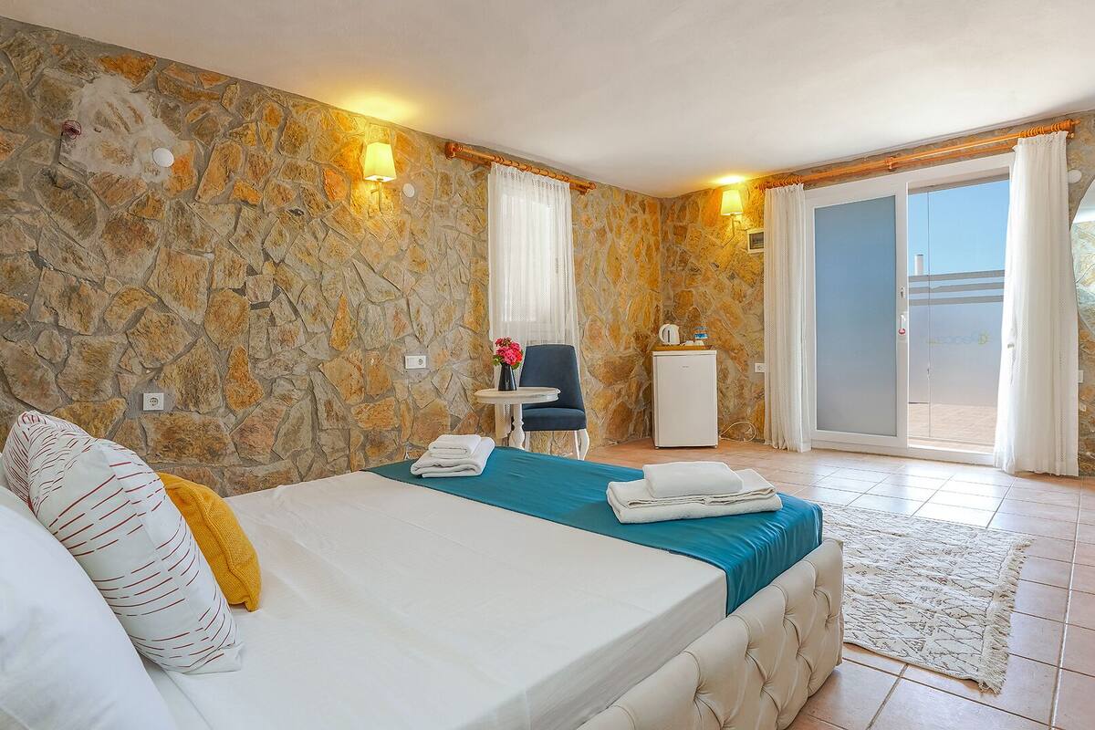 A hotel room with a stone wall, a bed with white bedding, a blue bedspread, a small table with a lamp, and a view through a glass door.