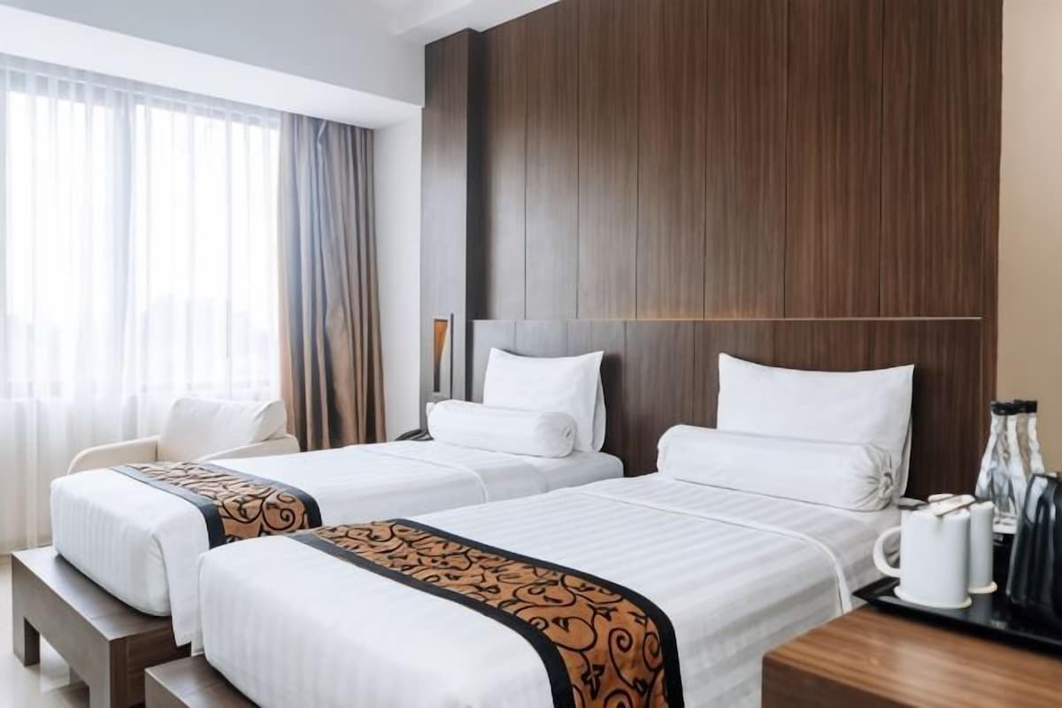 A hotel room with two beds, a wooden headboard, a bedside table with a coffee maker, and a window with curtains.