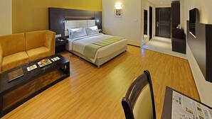 A hotel room with a bed, a sofa, a coffee table, a chair, and a desk with a newspaper. - Poppys Hotel (Madurai)