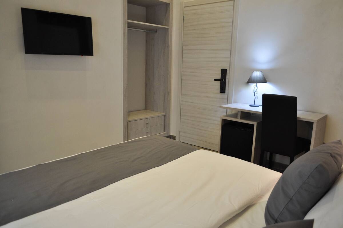 A bedroom with a bed, a desk with a chair, a wardrobe, and a TV mounted on the wall.