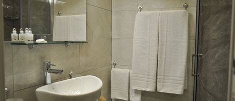 Un baño moderno con un lavamanos blanco grande, cabina de ducha de vidrio y dos toallas blancas colgadas en un perchero.