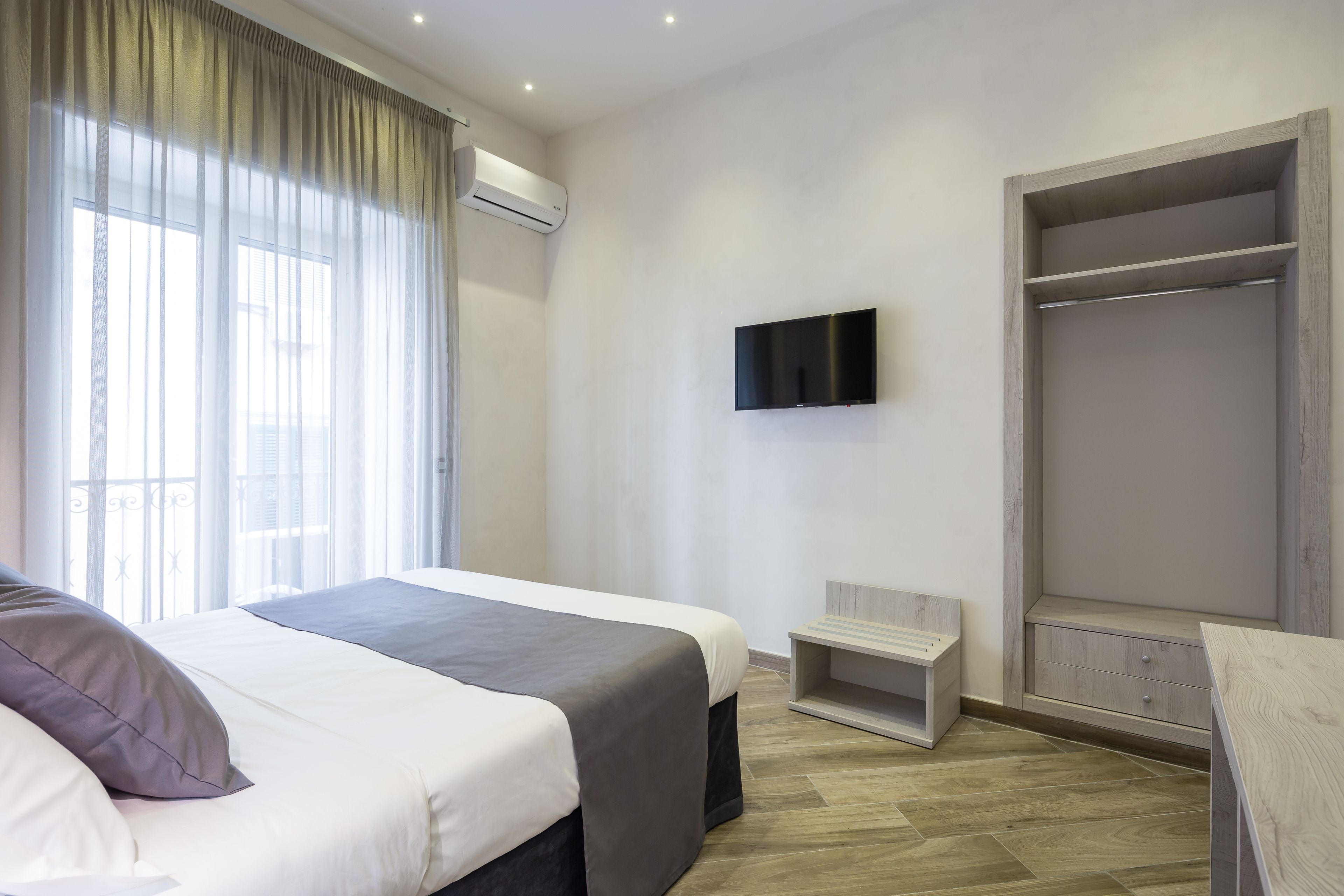 A hotel room with a bed, a TV mounted on the wall, a wardrobe, and a small seating area.
