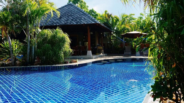 A swimming pool with a tiled edge, surrounded by lush greenery and a traditional building with a tiled roof.