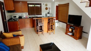 Un espacio habitable compacto con kitchenette, una zona de comedor con dos sillas de madera, un sofá con cojines y un televisor sobre un mueble de madera.