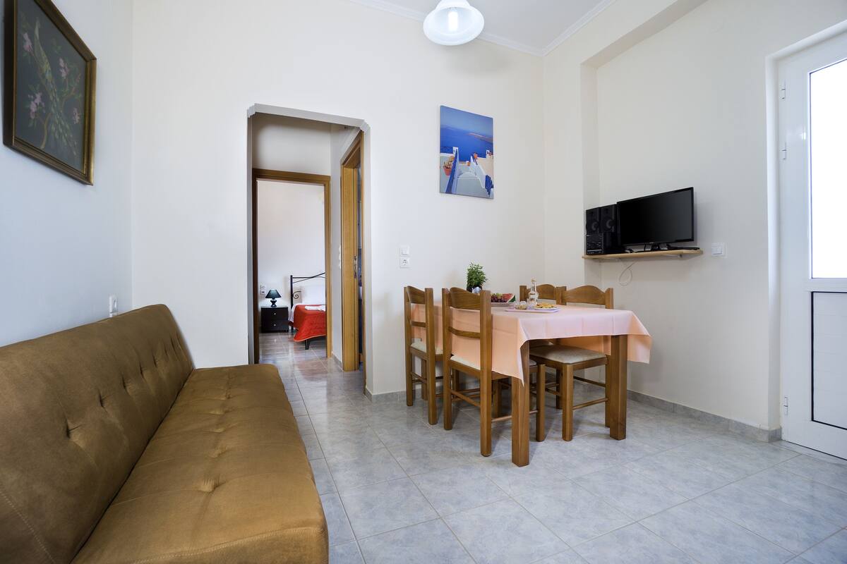 A living room with a brown sofa, a dining table with chairs, a TV on a shelf, and a painting on the wall.