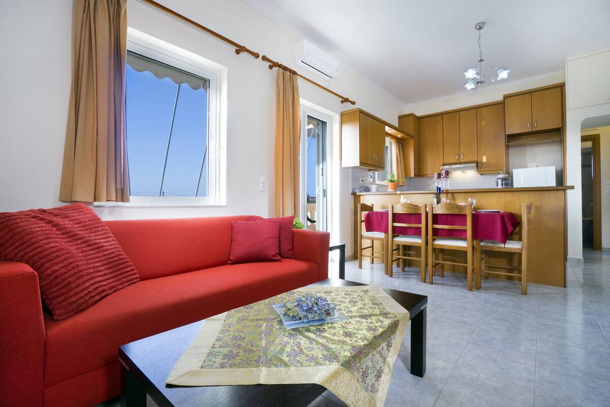 A compact living space with a red sofa, a dining area with wooden chairs, and a kitchen area with wooden cabinets.