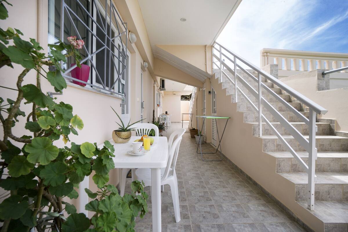 A clean, modern outdoor area with a white table set for dining, surrounded by potted plants. A staircase leads to an upper level, and a balcony is visible in the background.
