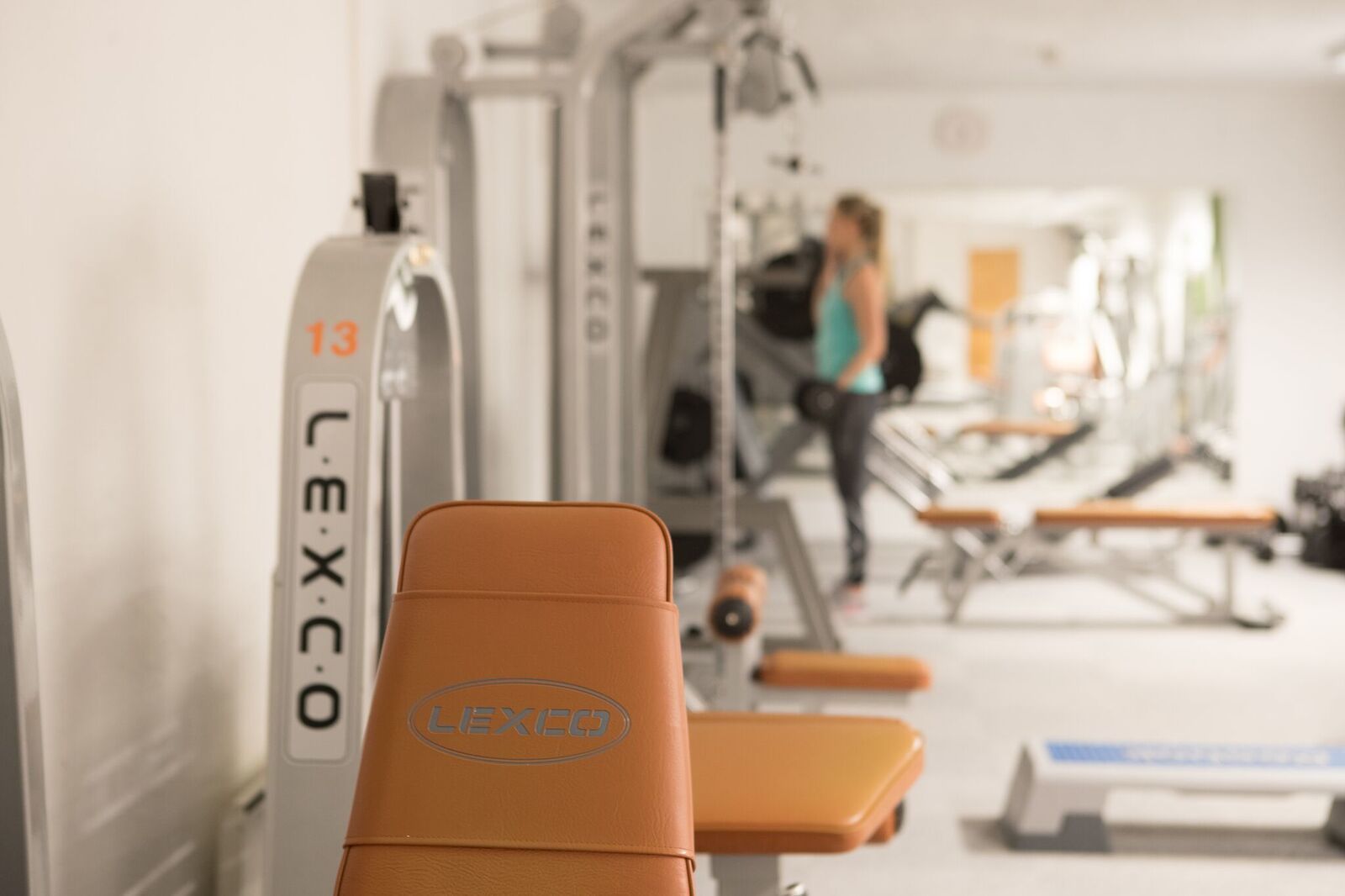 Un gimnasio con equipo de ejercicio, incluyendo un banco con la etiqueta "LIEXICO" y una máquina de pesas con el número "13".