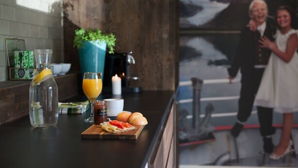 A kitchen counter with a glass bottle, a glass of orange juice, a jar of jam, and a plate with croissants and a red fruit slice.