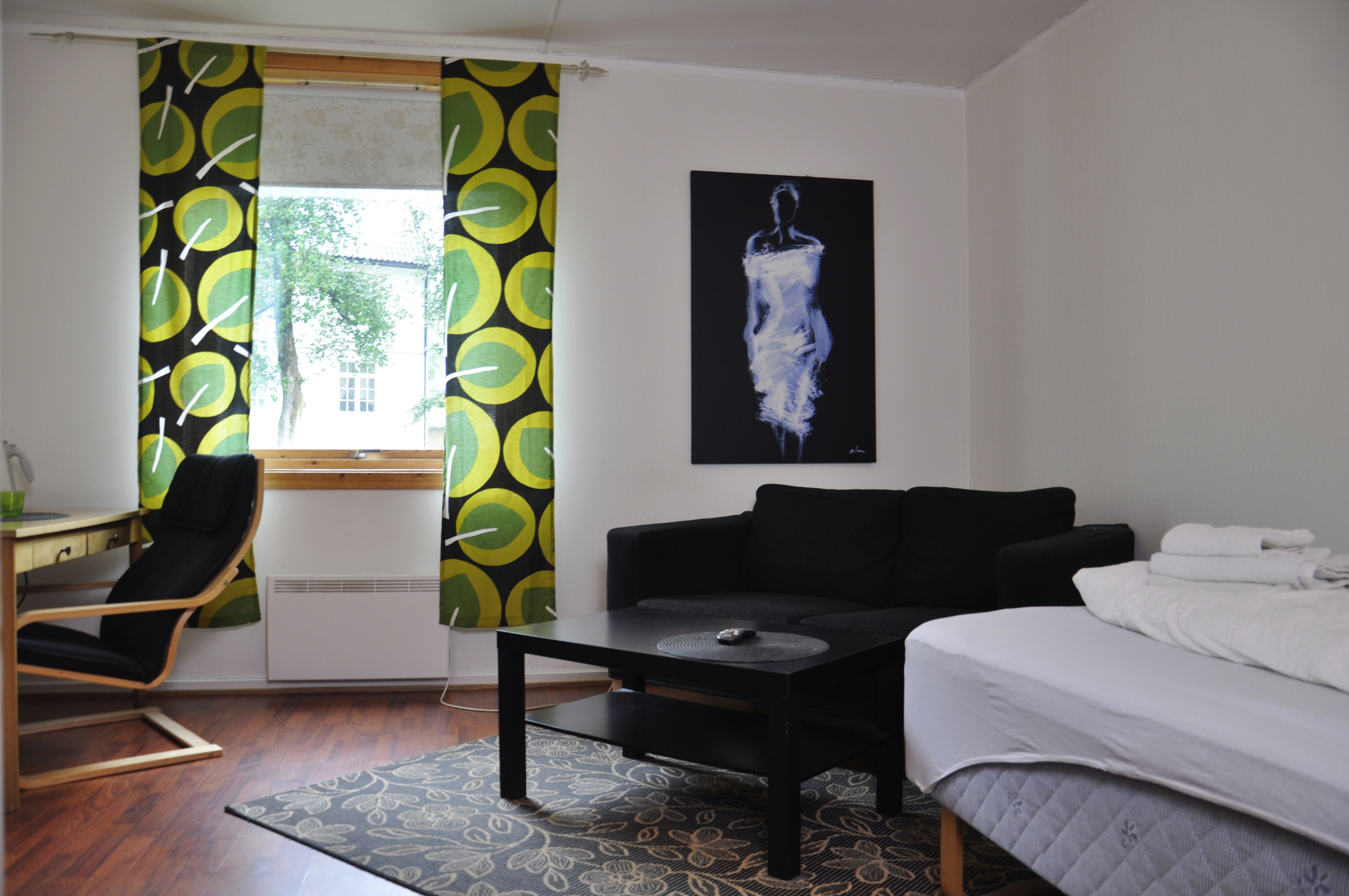Ett hotellrum med en säng, en svart soffa, ett litet bord, en stol och ett fönster med gardiner.