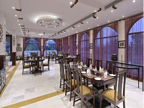A spacious dining area with multiple tables and chairs, large windows with curtains, and a modern ceiling design. - Talai Bagh Palace (Amer)