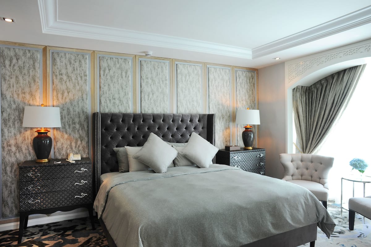 A bedroom with a tufted headboard, a large bed with grey bedding, two bedside tables with lamps, a chair, and a view of the outdoors through a window with curtains.