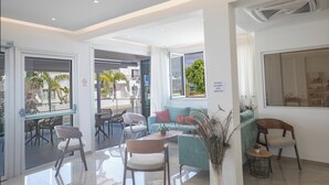 A modern hotel lobby with a seating area, a reception desk, and large windows offering views of the outdoors. - Pavlinia Hotel Apartments (Ayia Napa)