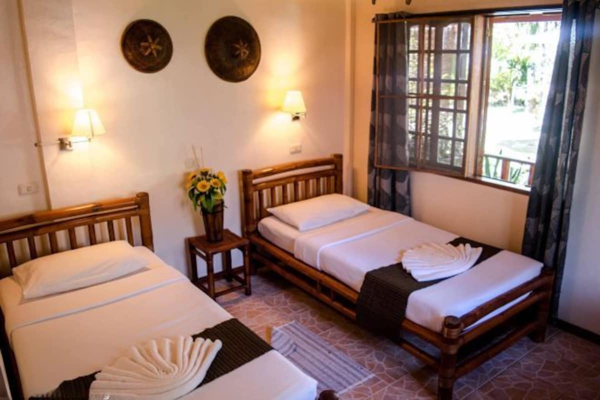 A room with two wooden beds, each with white linens and a small table beside them. There are wall-mounted lamps and a window with curtains.