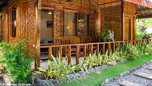 Edificio de madera tradicional con techo de paja, un área de estar al aire libre cubierta y un jardín bien cuidado con plantas tropicales.