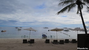 Una playa con sombrillas y sillas de descanso, barcos en el agua y una palmera.