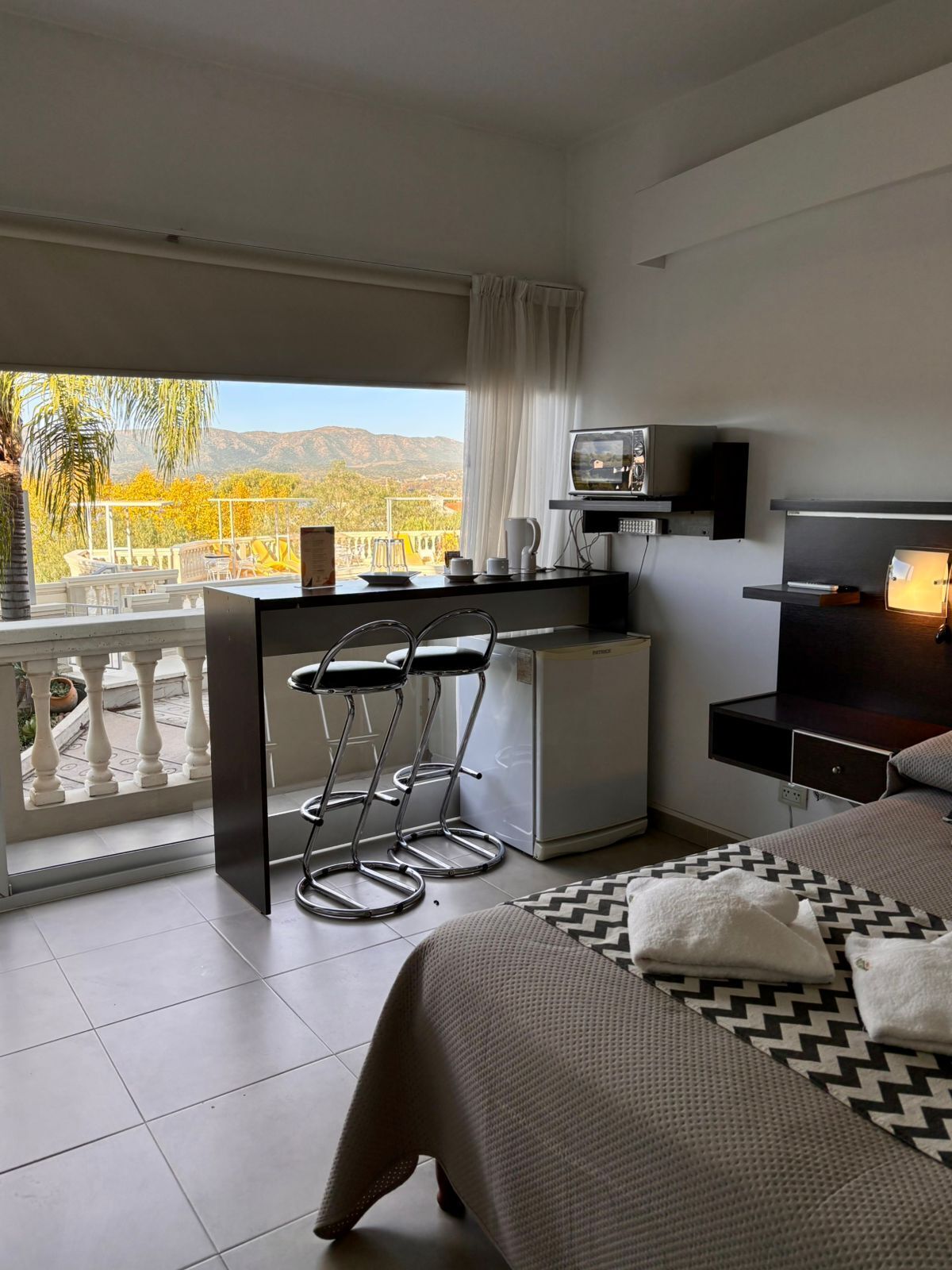 Un dormitorio moderno con cama, escritorio, silla, televisor y balcón con vistas a montañas.