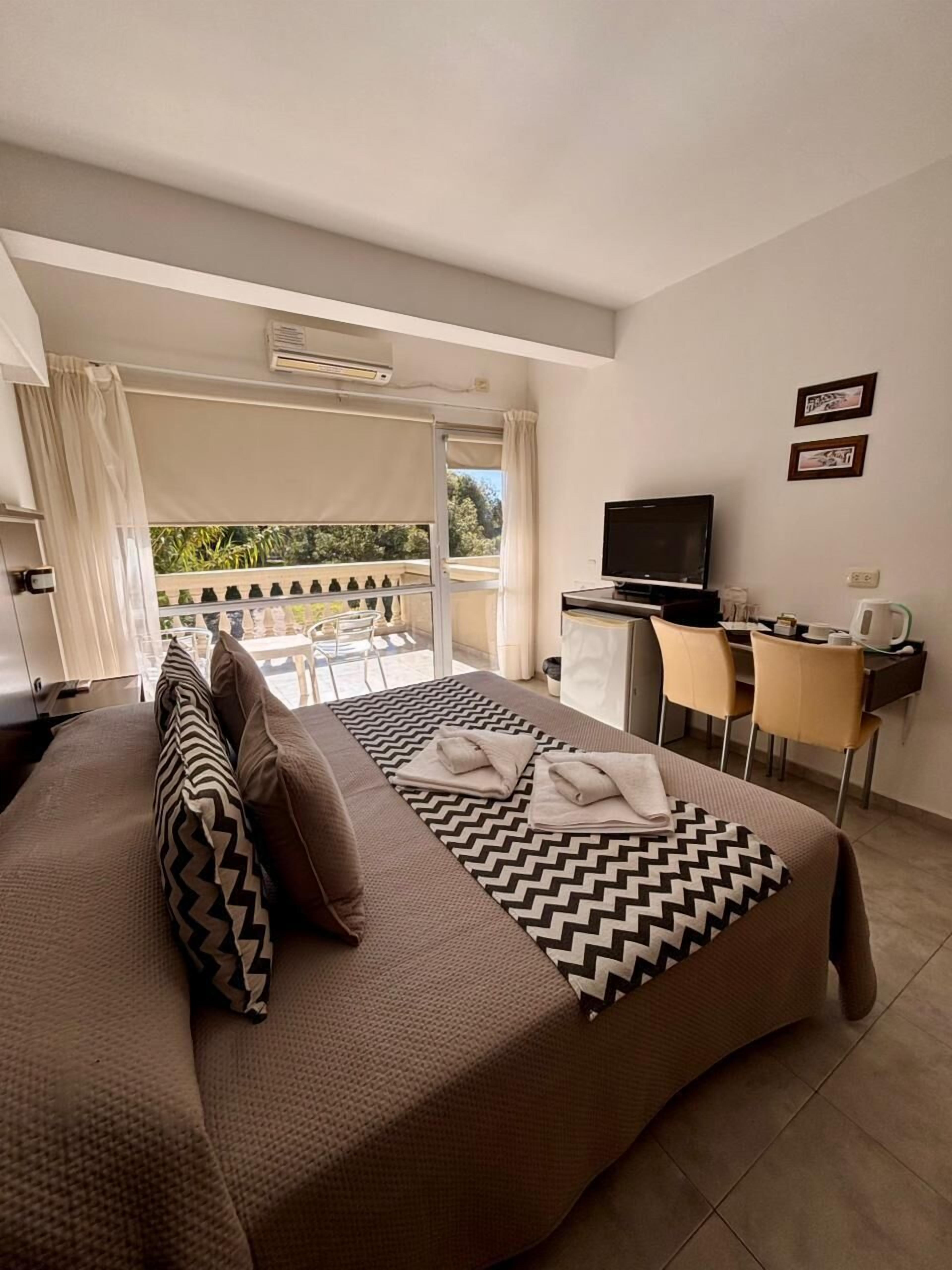 A hotel room with a bed, a desk, a TV, and a balcony with a view of greenery.