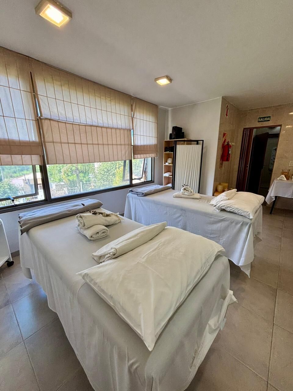 Een hotelkamer met twee bedden, een raam met zonwering, een kleine tafel met een lamp en een deur met een bordje.