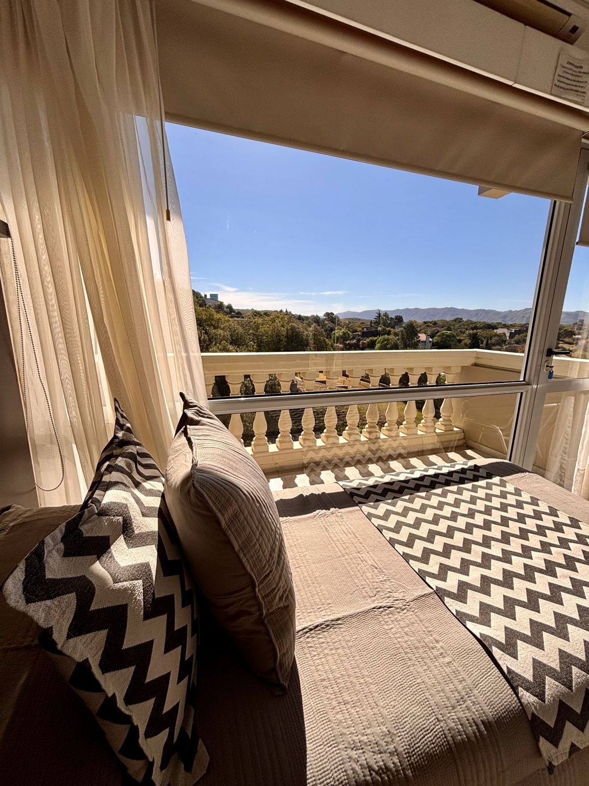 A balcony with a view of a mountainous landscape, a cushioned seating area with patterned cushions, and a window with sheer curtains.