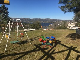 A playground with swings and colorful chairs, set against a backdrop of hills and a lake.