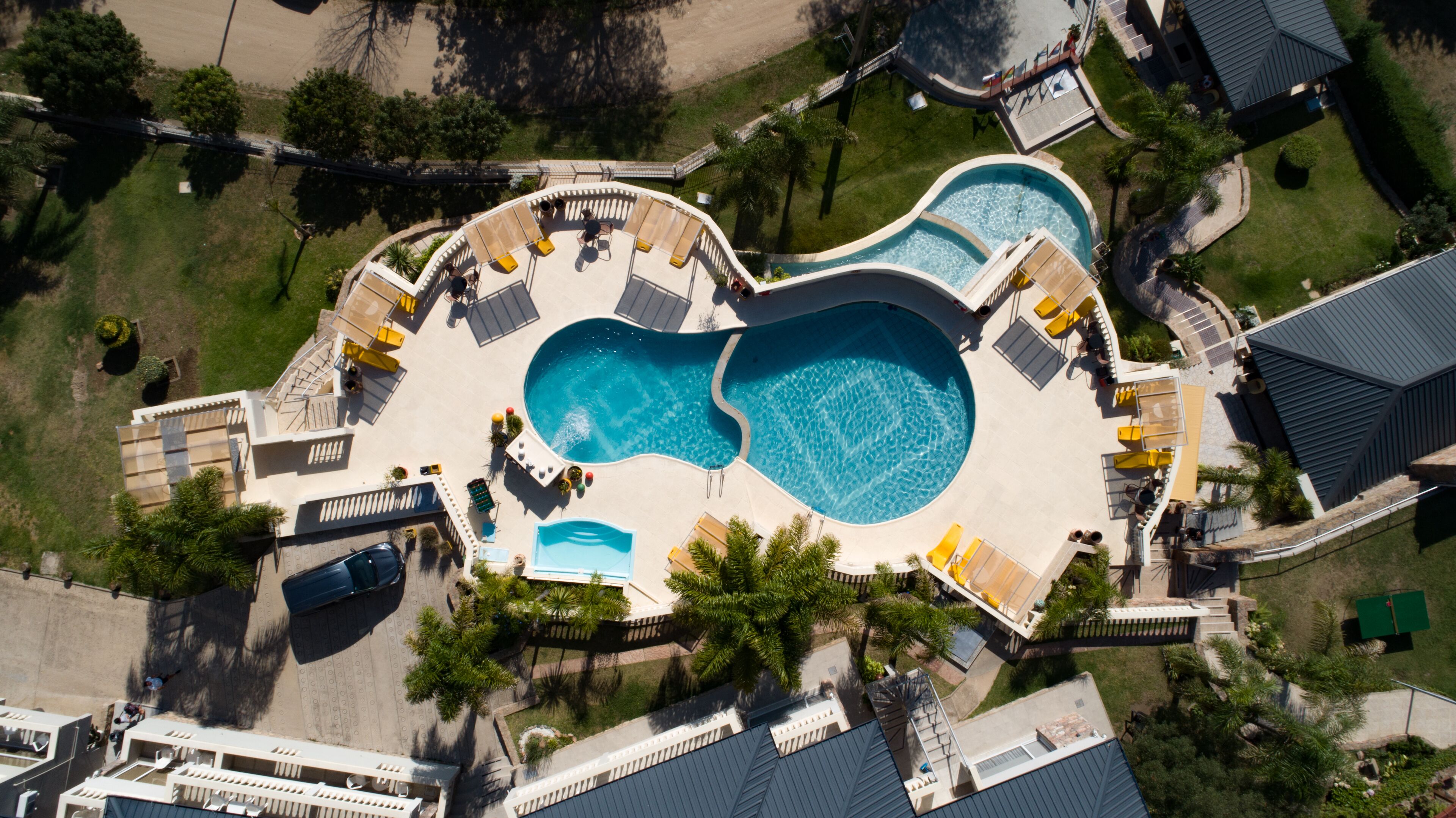 Vista aérea de un complejo turístico con una gran piscina central, varias piscinas más pequeñas y tumbonas dispuestas alrededor de las piscinas.