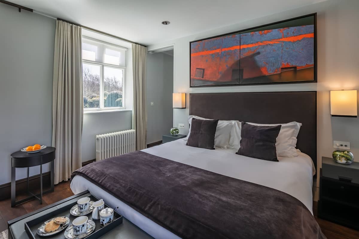 A modern hotel room with a large bed, a painting above the headboard, a window with curtains, a small table with a tray of breakfast, and a radiator.
