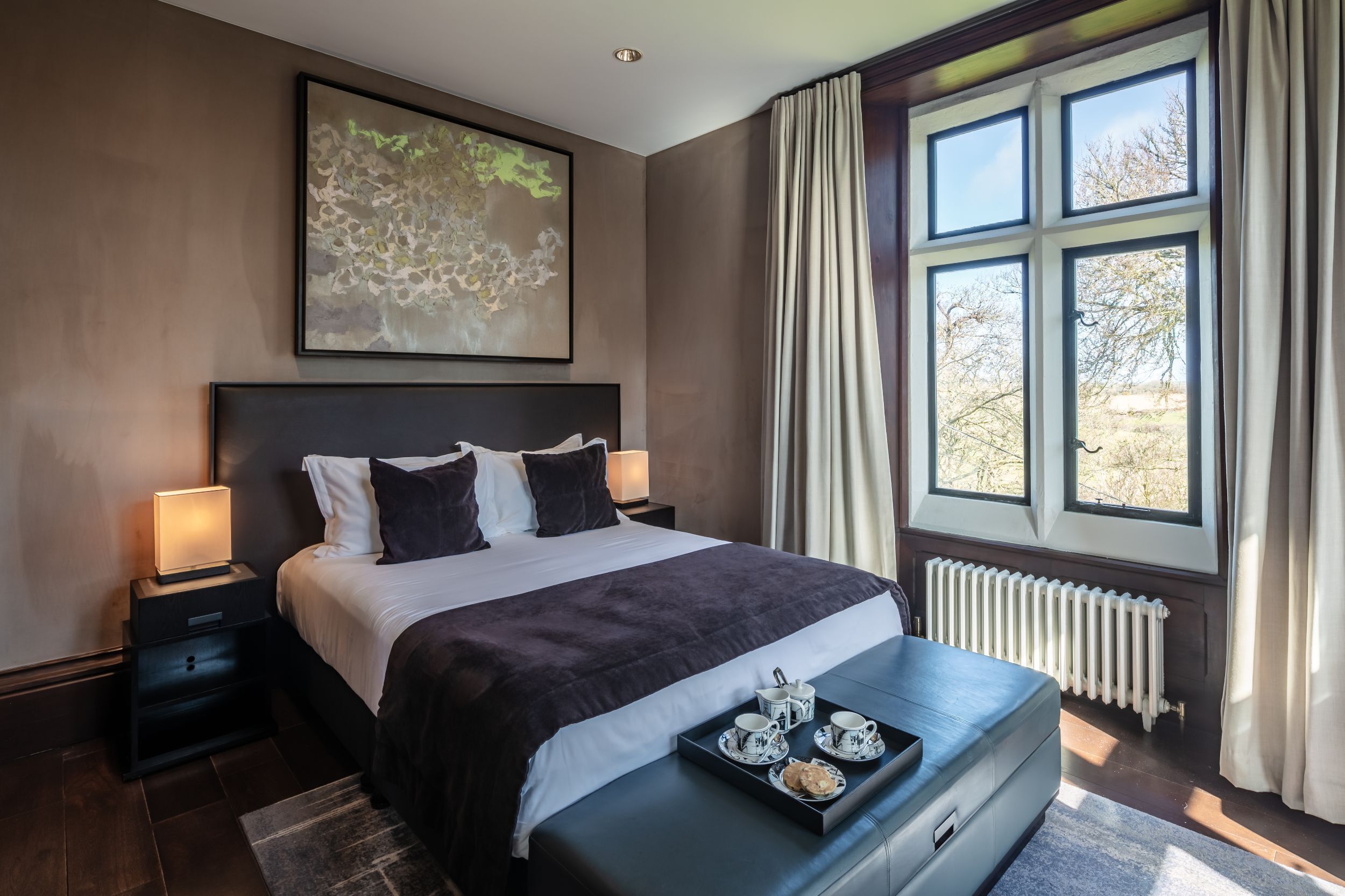 A modern hotel room with a large bed, a bedside table with a lamp, a view of trees through the window, and a tray with cups and pastries on the bench.
