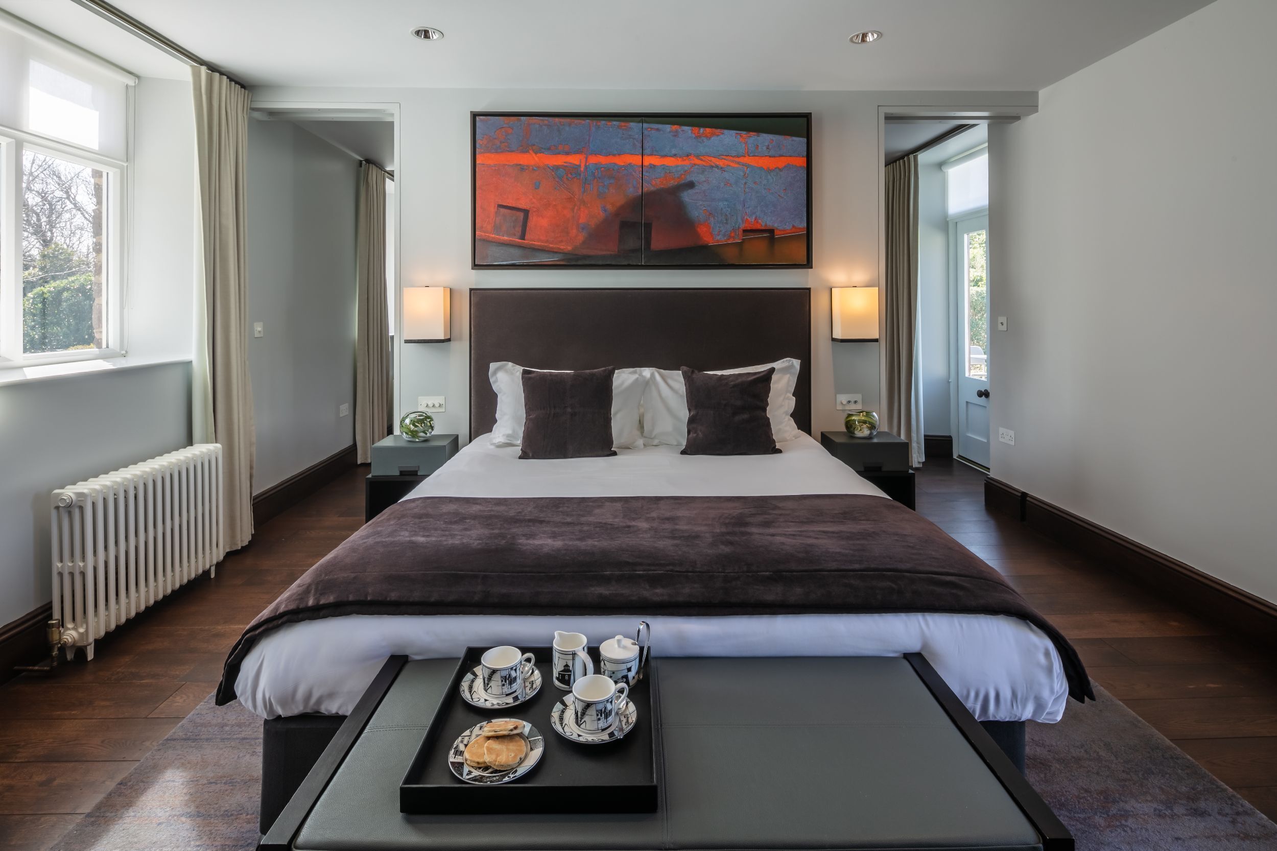 A modern hotel room with a large bed, bedside tables, a tray with breakfast items, and a painting on the wall.