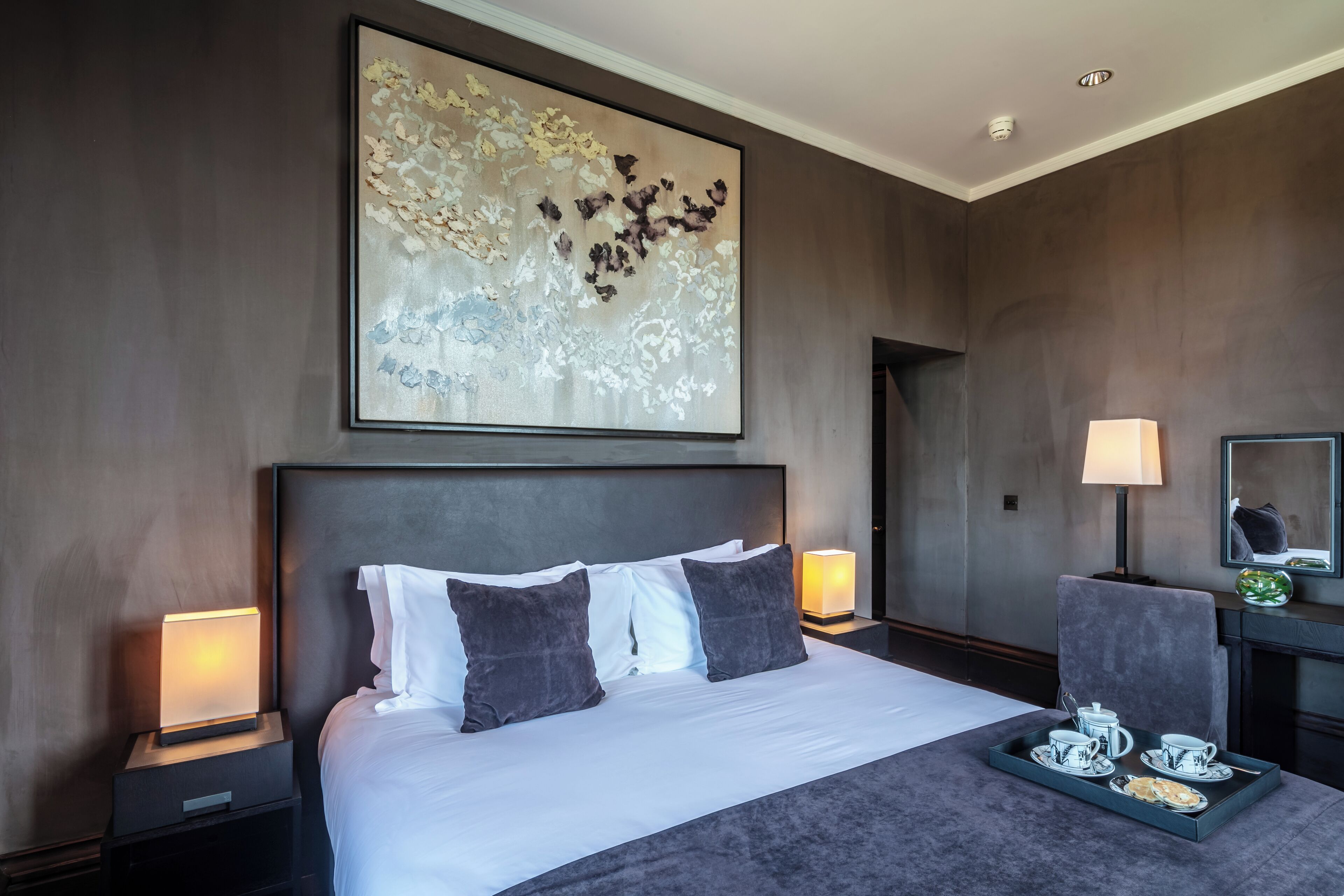 Habitación de hotel con una cama grande, dos mesitas de noche con lámparas, una bandeja con té y pasteles, y un cuadro abstracto de gran tamaño en la pared.