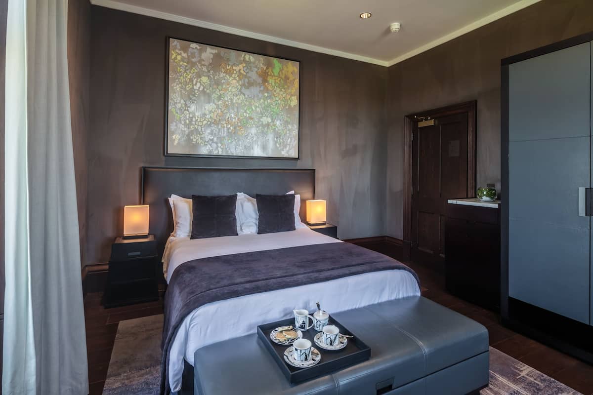 A modern hotel room with a large bed, bedside lamps, a tray with tea set, and a large abstract painting on the wall.