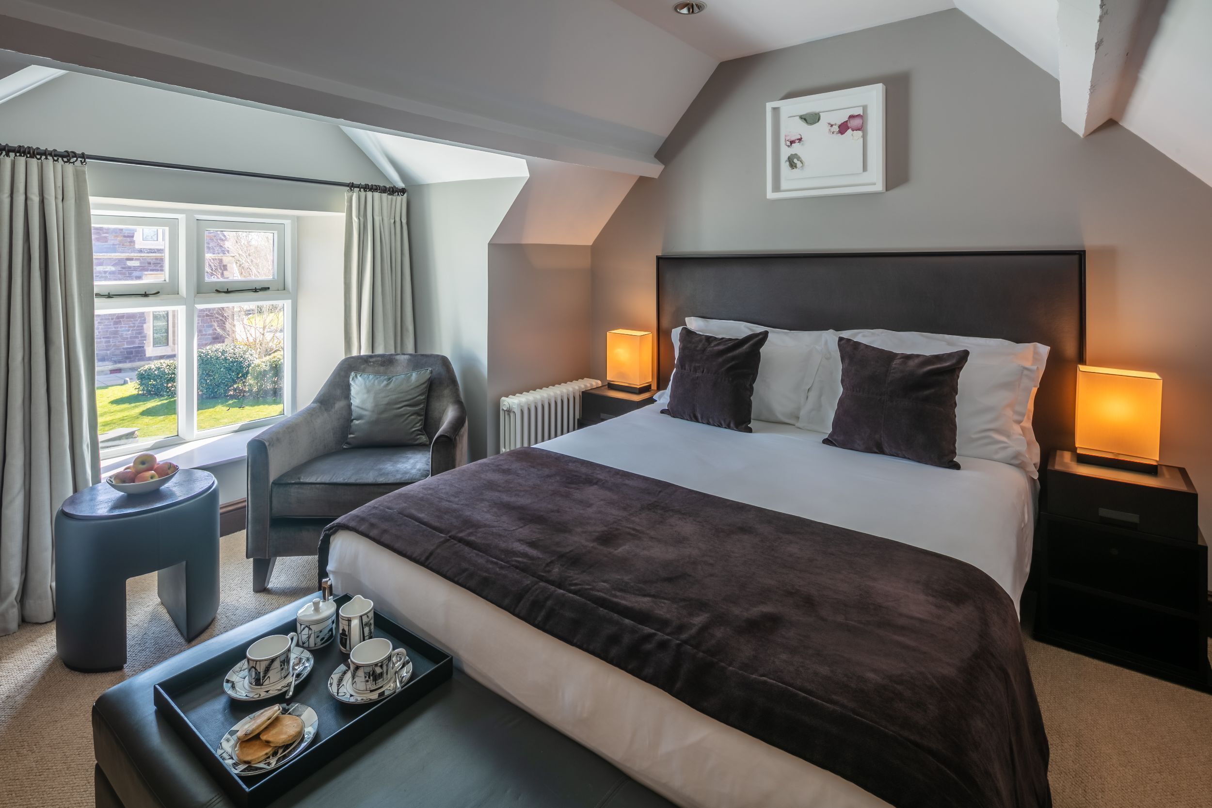A bedroom with a large bed, a sitting area with a chair, a small table with a tray of breakfast, and a view of the outdoors through a window.