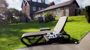 Un edificio de piedra con techo inclinado y un césped. Hay dos sillones de exterior con una mesita de bandeja, y un juego de tazas blancas y una tetera.