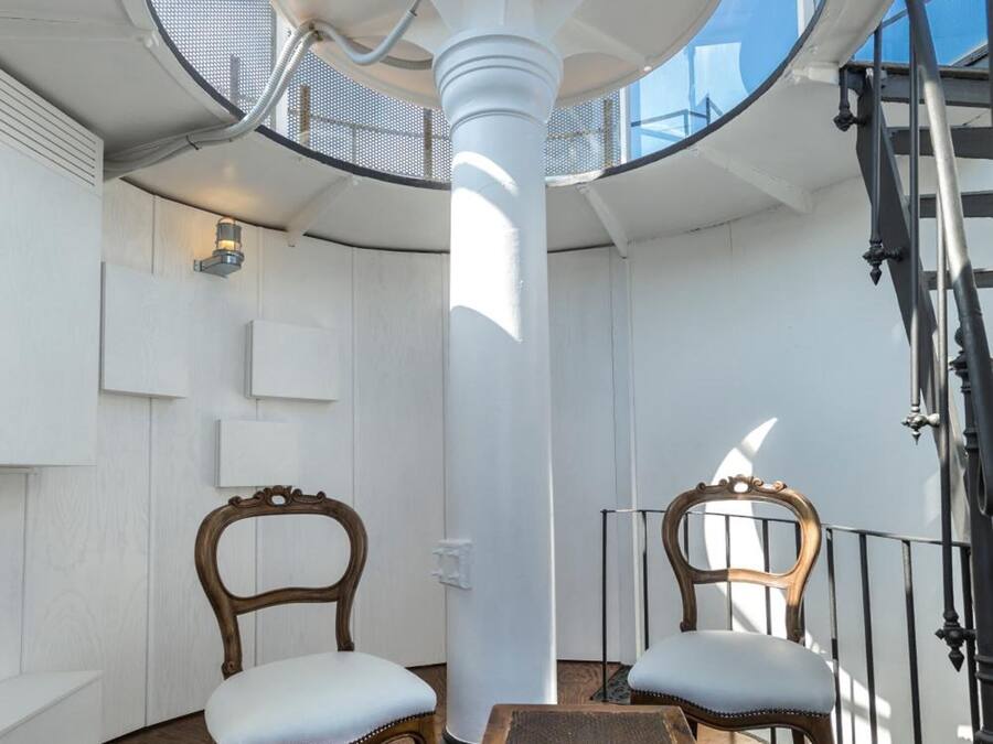 Une pièce circulaire avec une colonne centrale, deux chaises en bois avec des coussins, une petite table en bois et un escalier sur le côté droit.