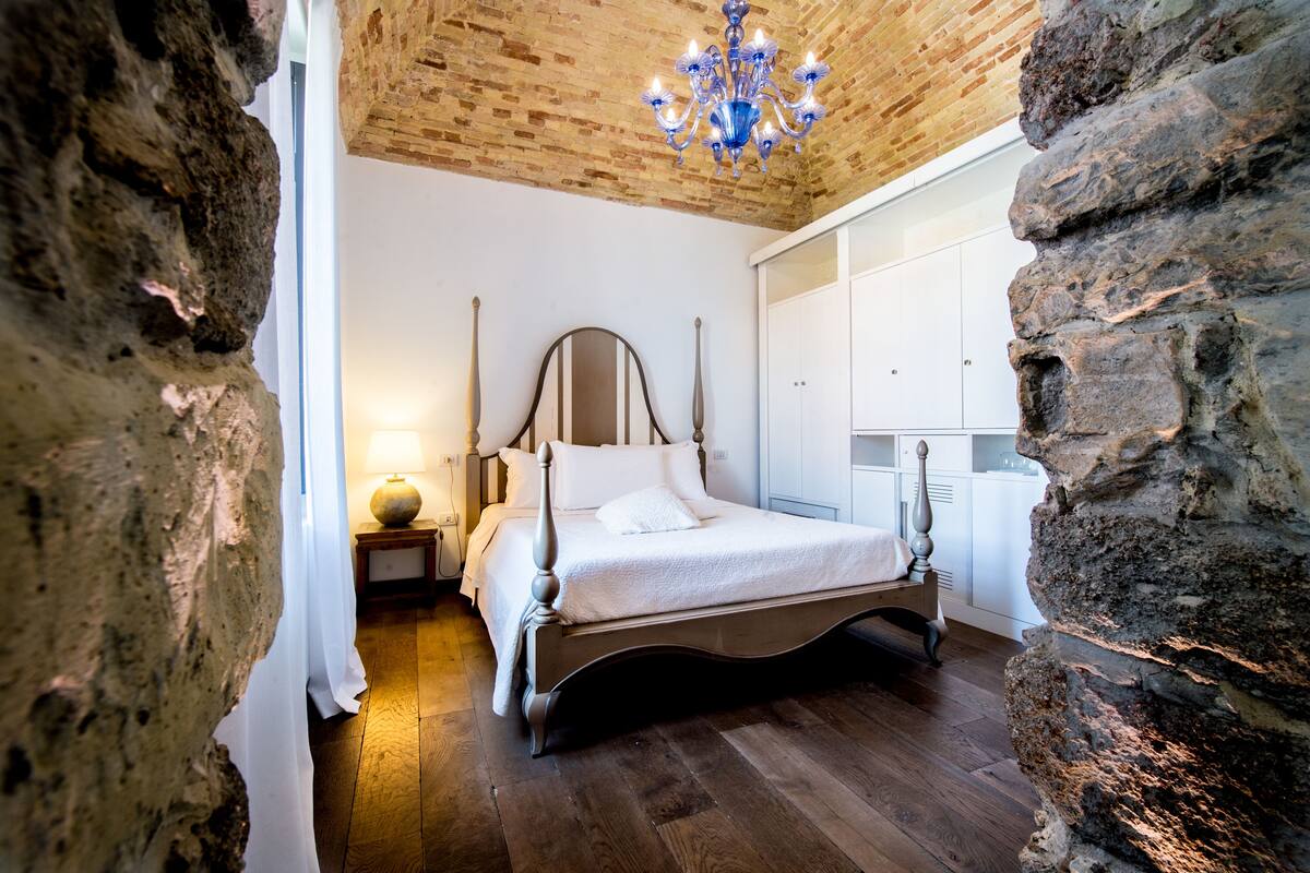 A bedroom with a wooden bed, a nightstand, a lamp, and a stone wall.