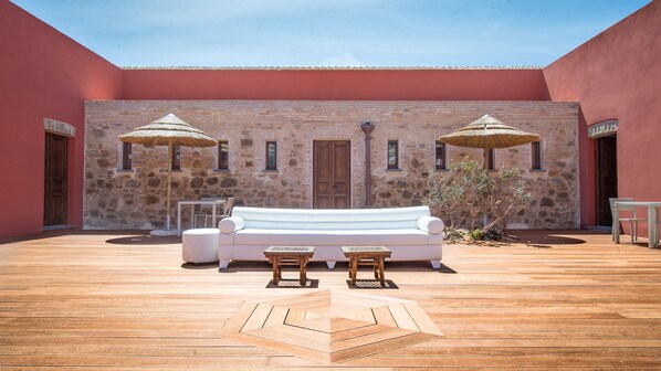 Une cour avec un mur en pierre, un canapé blanc, deux petites tables et deux parasols à toits de chaume.
