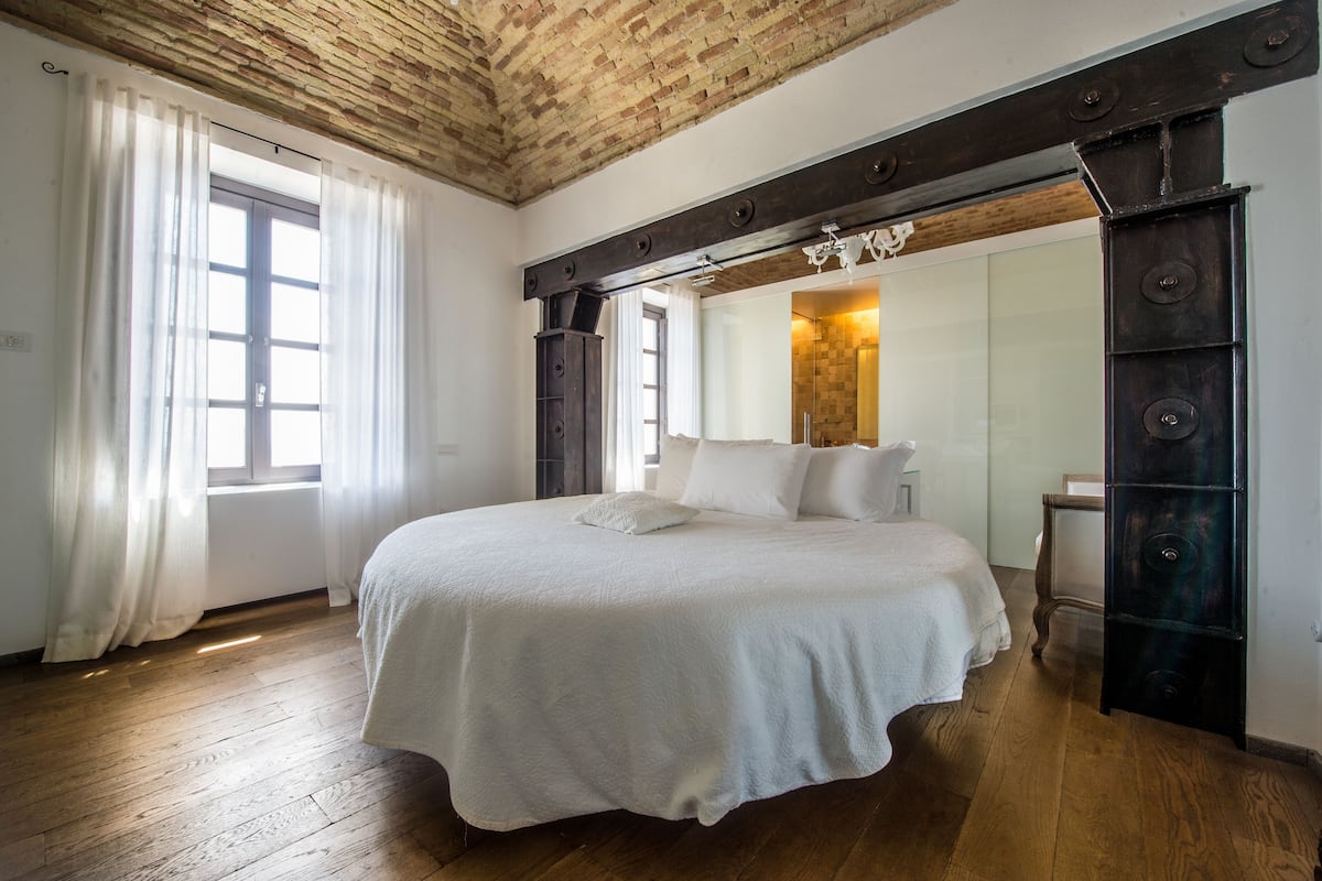 A bedroom with a large bed, wooden flooring, a brick ceiling, and a large mirror above the bed.