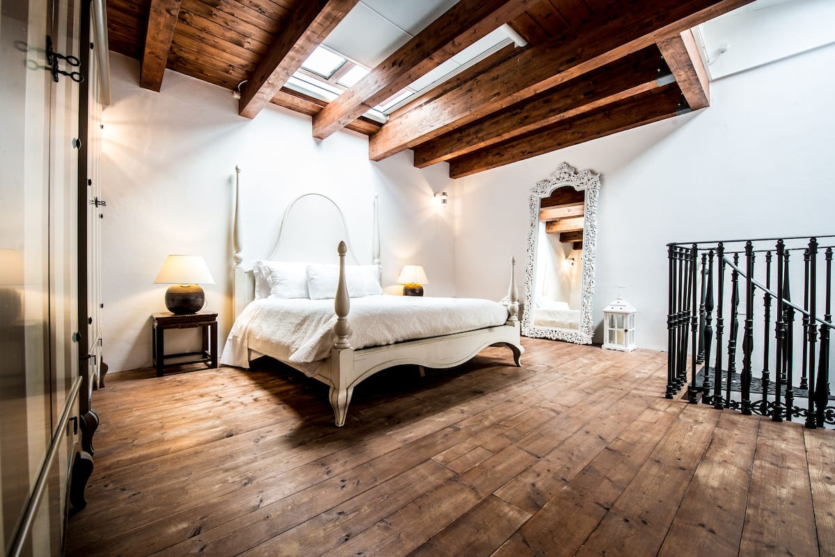A bedroom with a wooden floor, a bed with a white headboard, a small table with a lamp, and a black railing staircase.