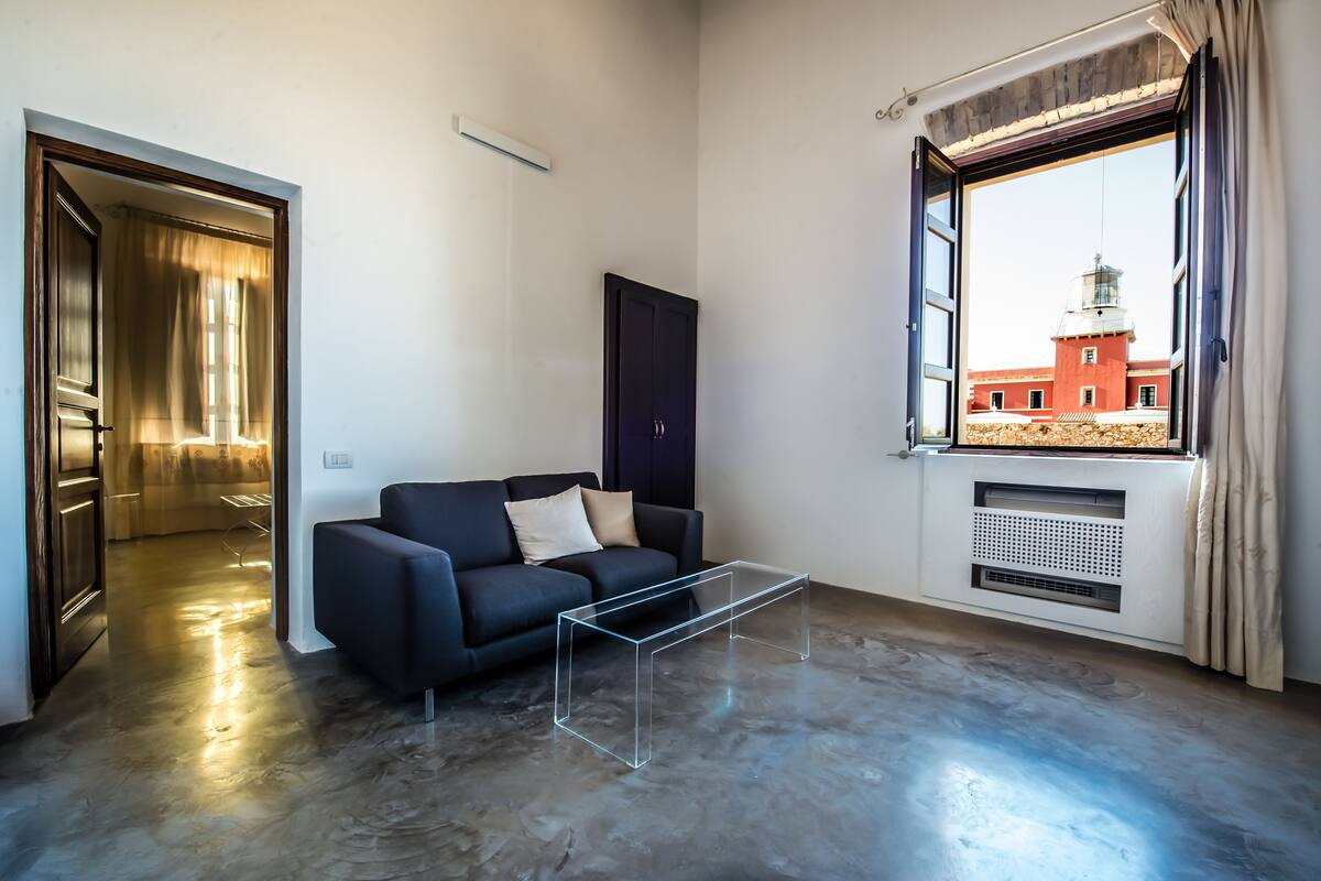 A modern living room with a dark sofa, a glass coffee table, and a view of a red building through a window.