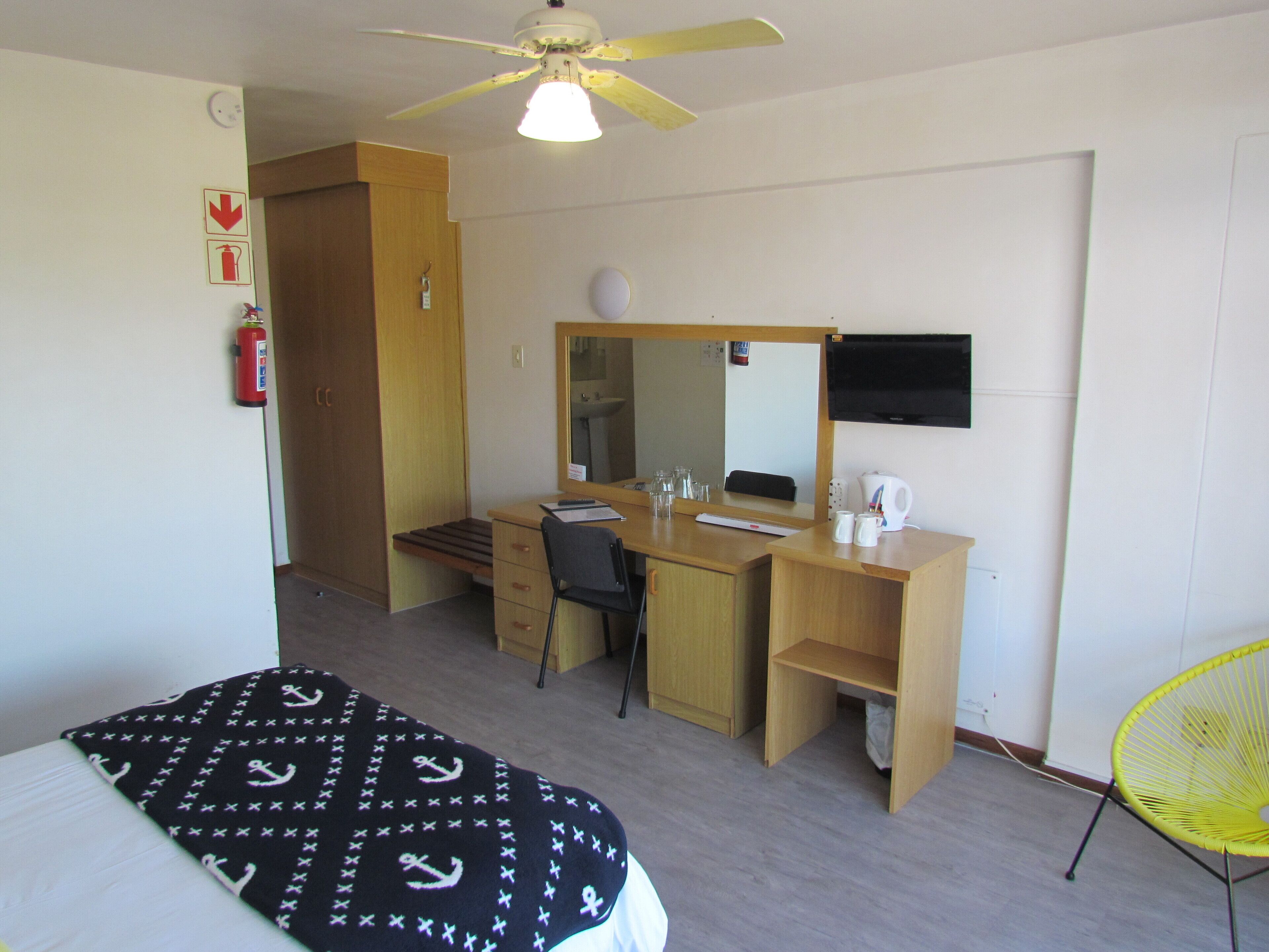 Habitación de hotel con cama, escritorio con silla, espejo, ventilador de techo y extintor.