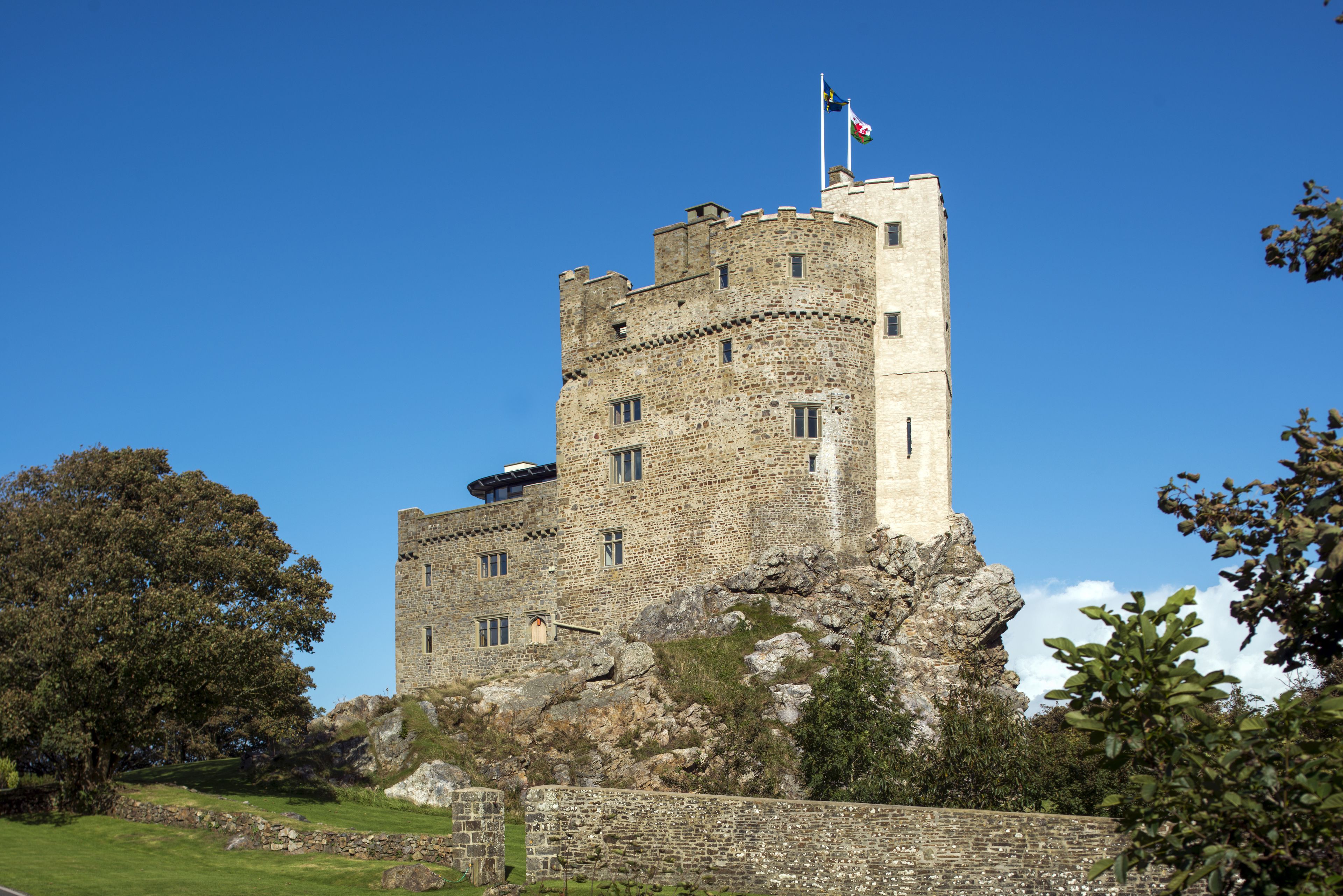 A historic stone castle with a flag on top, surrounded by greenery and a stone wall.