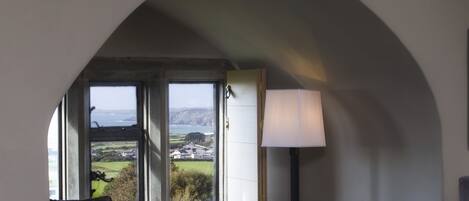 A room with a large arched window, a sofa, a table with a lamp, and a view of a landscape.