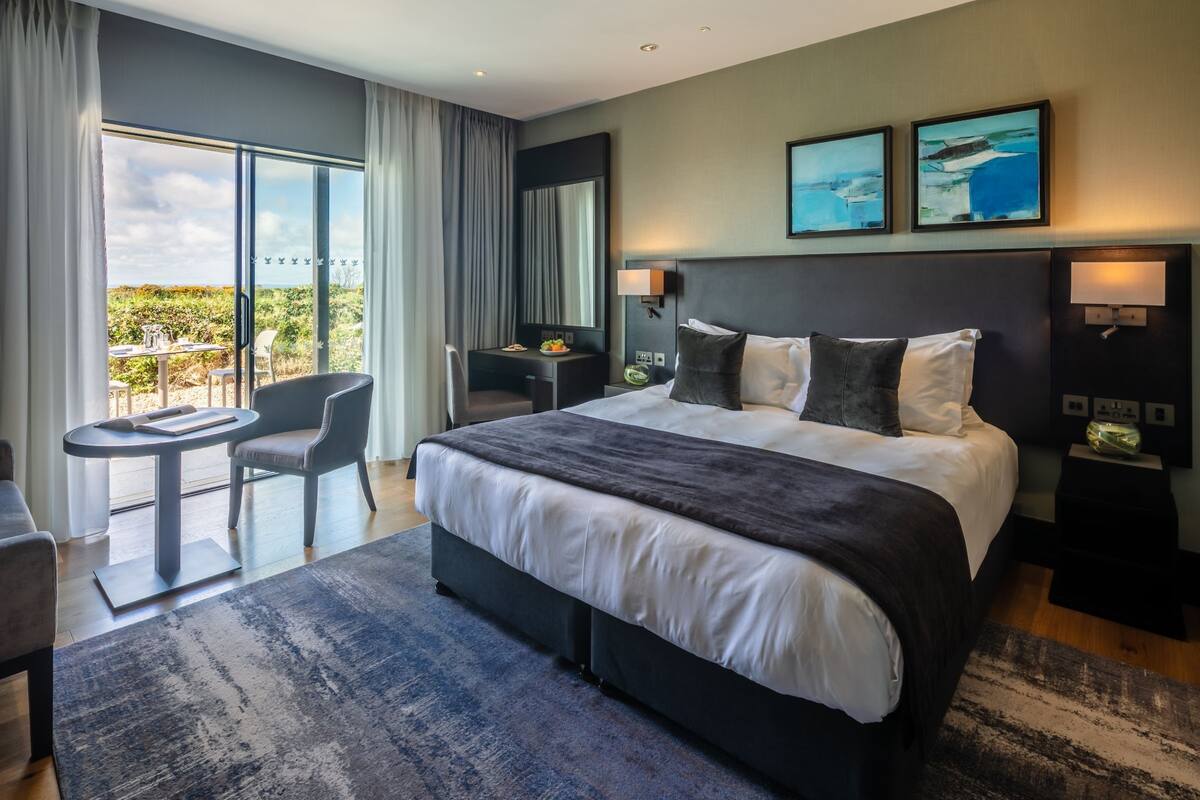 A modern hotel room with a large bed, a desk, a chair, and a balcony with a view.